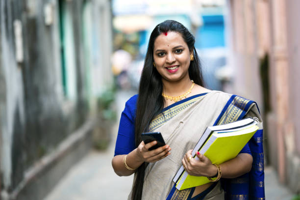 Outdoor image of Young Indian female School Teacher or College Professor using smartphone and holding books in hand.